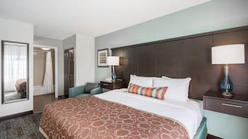 Quarto de hotel com uma cama grande, mesa de cabeceira, luminária, cadeira, banheiro com chuveiro e espelho.