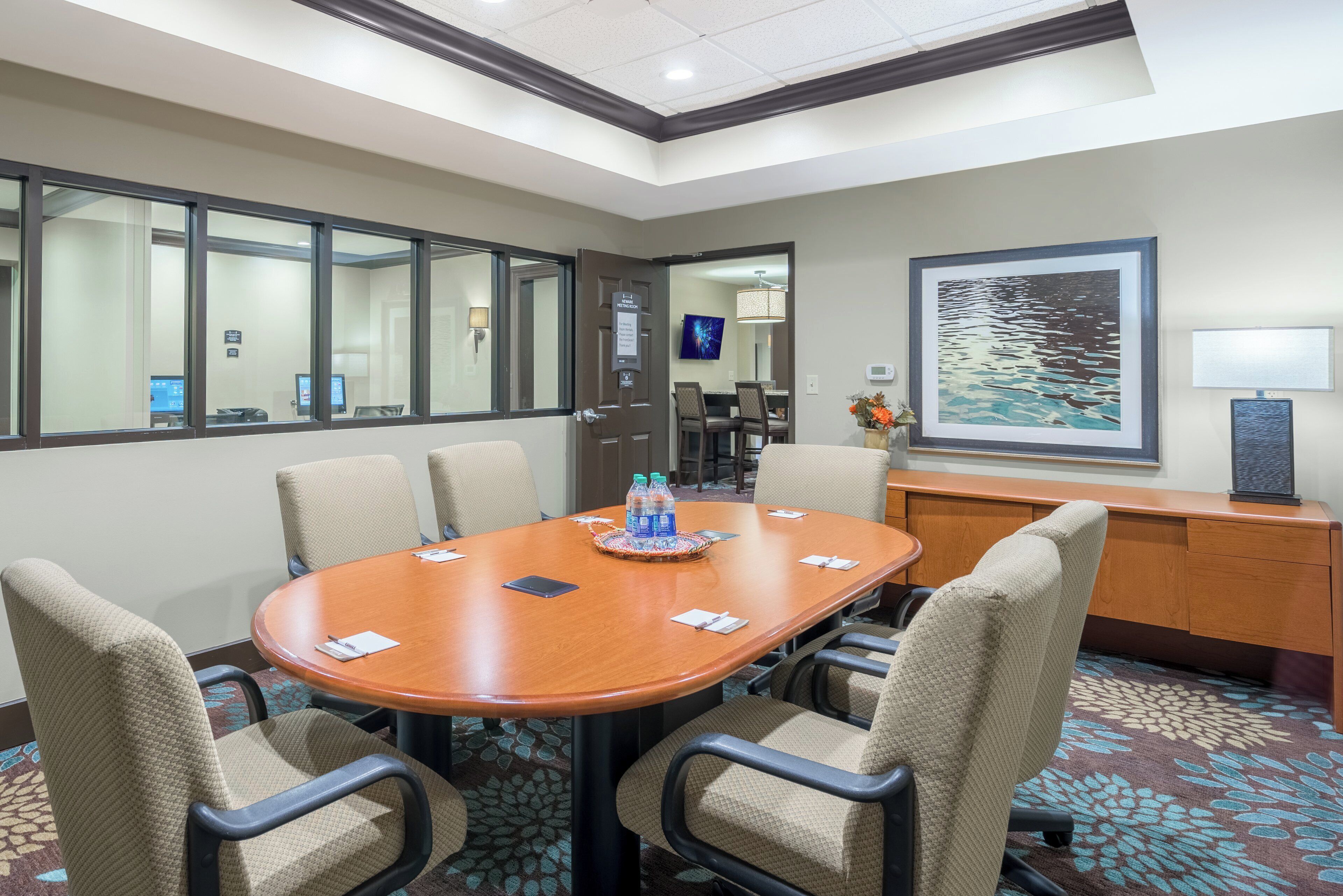 A conference room with a wooden table, six chairs, a water dispenser, and a painting on the wall.