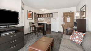 A compact living space with a kitchenette, a flat-screen TV, a sofa with a polka dot pillow, and a bench. - Staybridge Suites Wilmington-Newark by IHG (Newark)