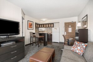 Een ruimtelijk ingerichte woonruimte met een kitchenette, een flatscreen televisie, een bank met een stippen kussen en een bankje.