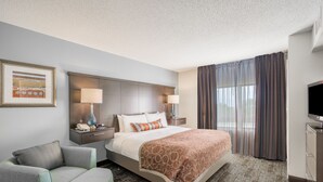 A hotel room with a bed, two bedside tables with lamps, a chair, a TV, and a large window with curtains. - Staybridge Suites Wilmington-Newark by IHG (Newark)