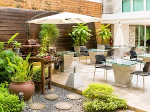 Een terras buiten met glazen tafels en stoelen, omgeven door potplanten en een bakstenen muur.