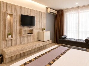 A modern hotel room with a wooden accent wall, a flat-screen TV, a small cabinet, a sofa, and a large window with curtains. - Swana Bangkok Hotel (Bangkok)