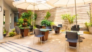 An outdoor patio area with multiple tables and chairs, some under umbrellas, surrounded by potted plants and a wooden wall. - Swana Bangkok Hotel (Bangkok)