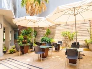 An outdoor patio area with multiple tables and chairs, some under umbrellas, surrounded by potted plants and a wooden wall.