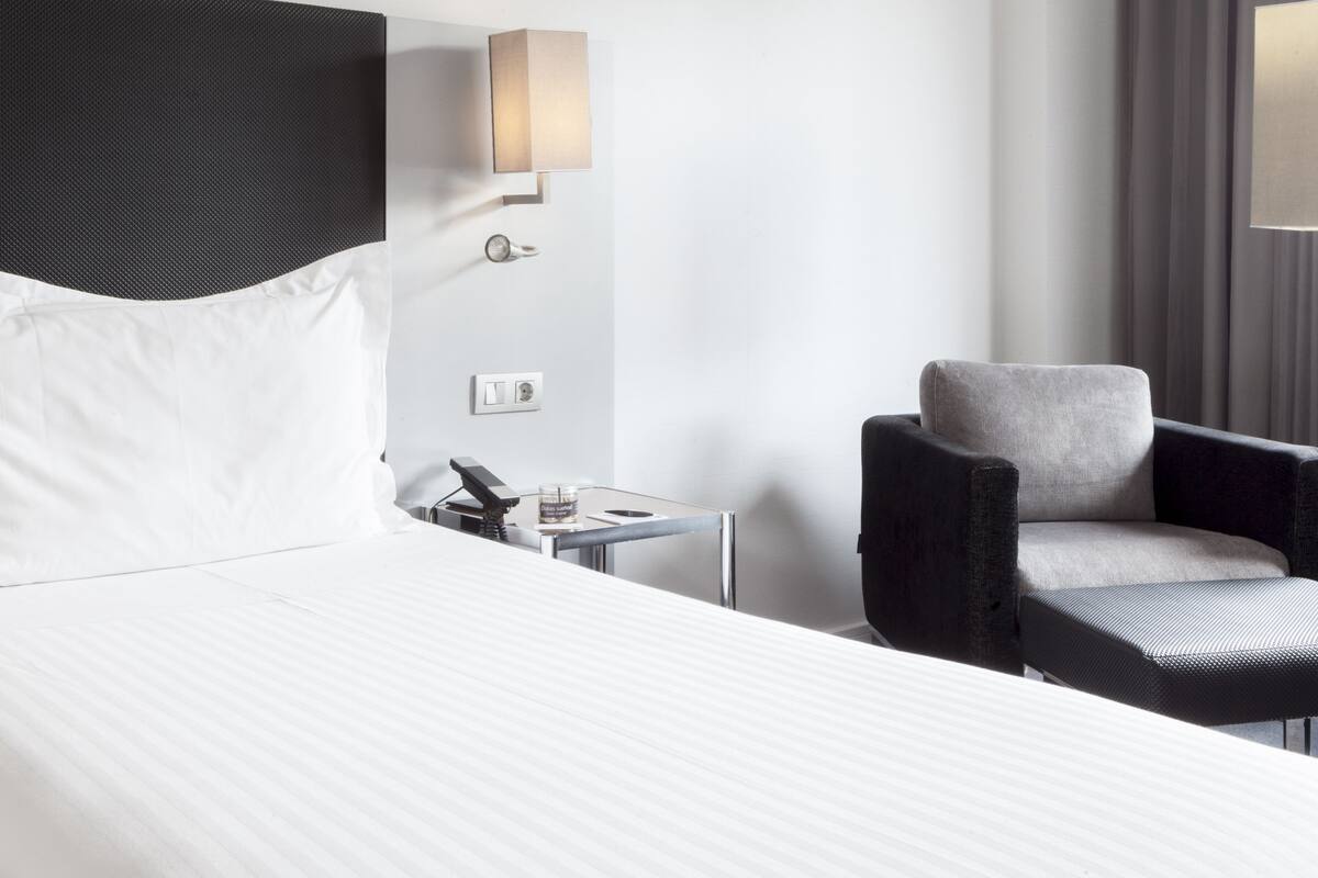 Une chambre d’hôtel comprenant un lit, une chaise, une table de chevet avec une lampe et un luminaire fixé au mur.