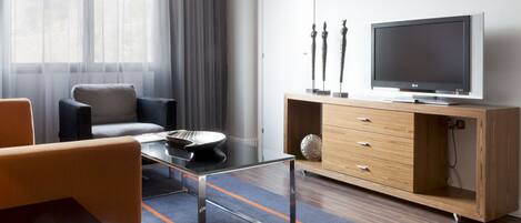 A modern living room with a glass coffee table, a sofa, a TV on a wooden cabinet, and a view through a window with sheer curtains.