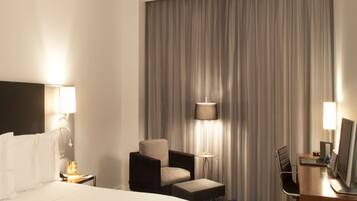 A hotel room with a large bed, a desk with a computer, a chair, a desk lamp, and a window with curtains.