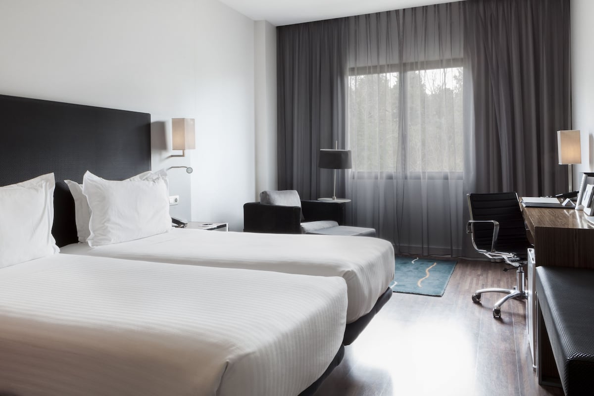 Une chambre d’hôtel avec deux lits, un bureau, une chaise et une grande fenêtre avec des rideaux.