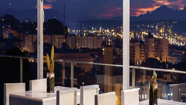A rooftop terrace with white tables and chairs, a decorative golden statue, and a cityscape view at dusk.