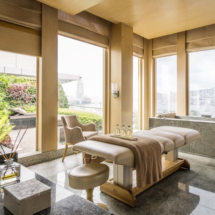 A modern spa room with a massage table, two chairs, a large window with a city view, and a small sink.
