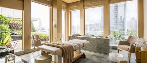 A modern spa room with a massage table, two chairs, a large window with a city view, and a small sink.