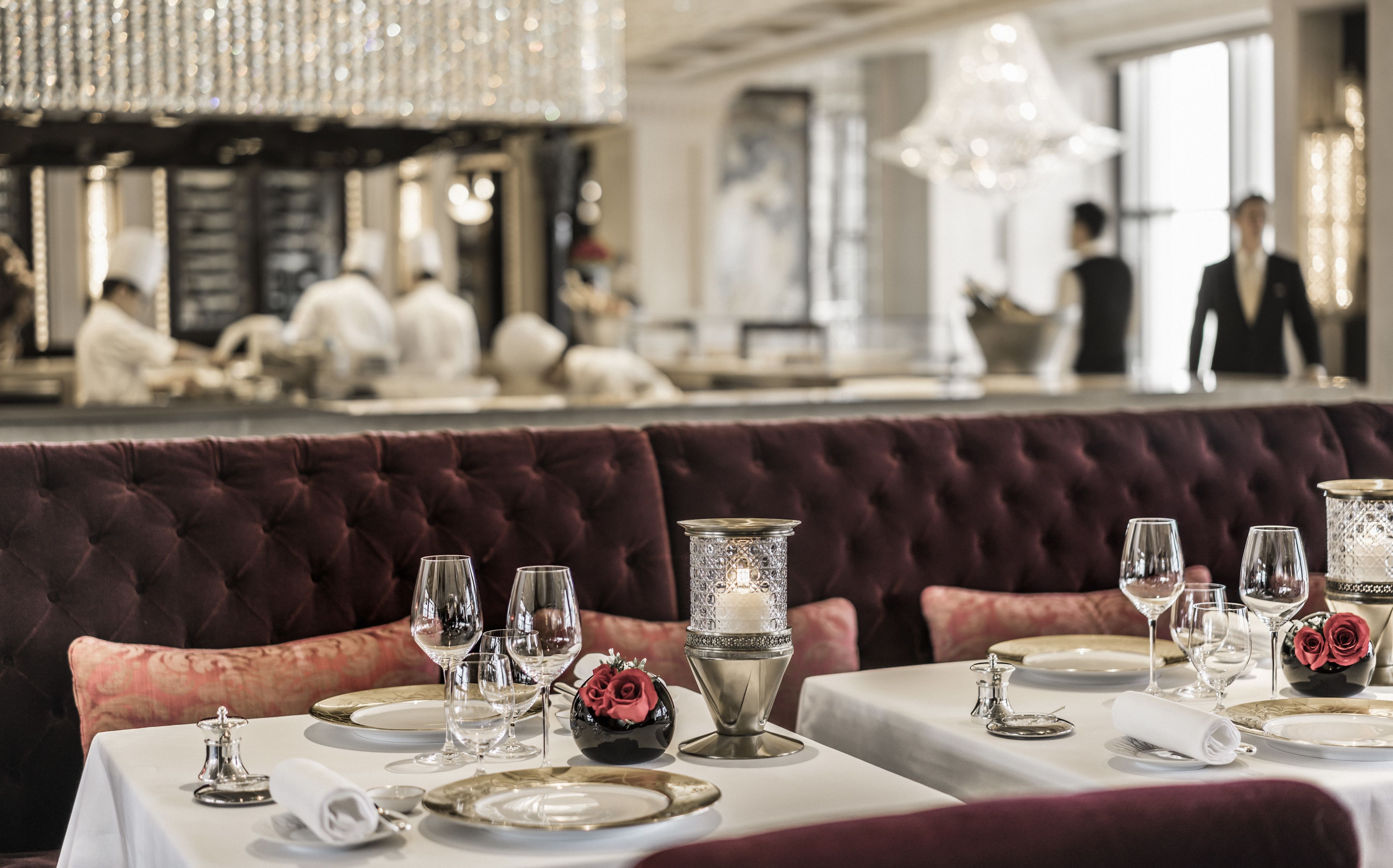 A dining area with tables set for a meal, featuring a plush red upholstered booth, elegant table settings, and a chandelier overhead.