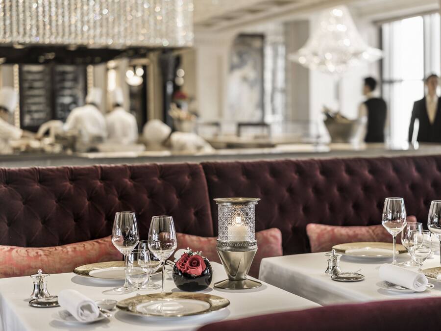 A dining area with tables set for a meal, featuring a plush red upholstered booth, elegant table settings, and a chandelier overhead.