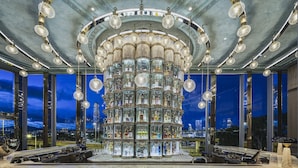 A bar area with a central display of liquor bottles, overhead chandeliers, and a city view through large windows.