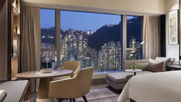 A modern hotel room with a large window offering a city view, a sofa, a dining table, and a bed.