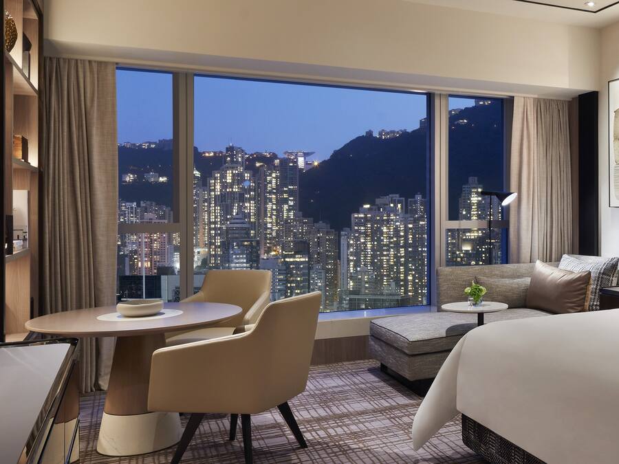 A modern hotel room with a large window offering a city view, a sofa, a dining table, and a bed.