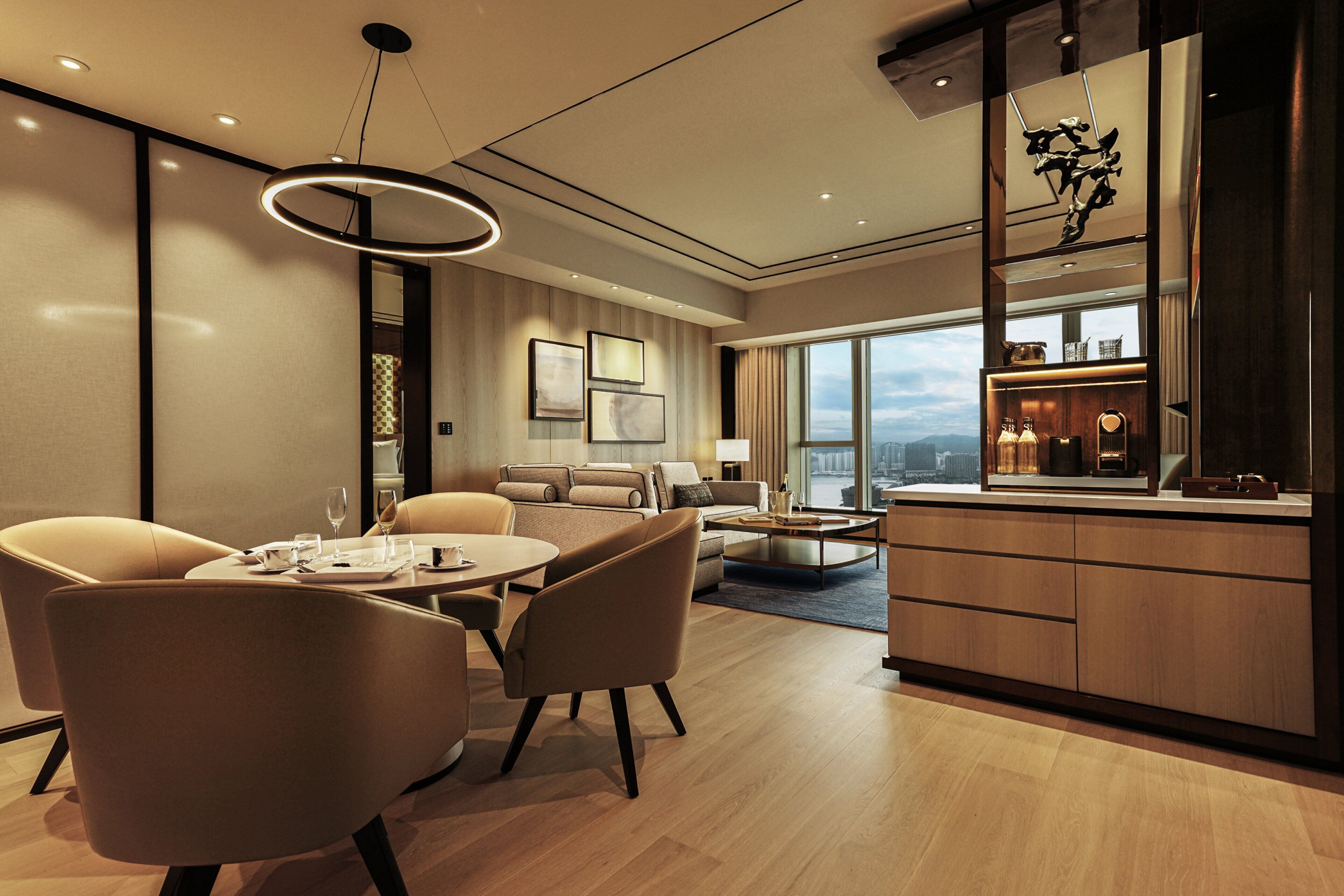 A modern hotel room with a dining area, a sofa, a coffee table, and a view of the city.