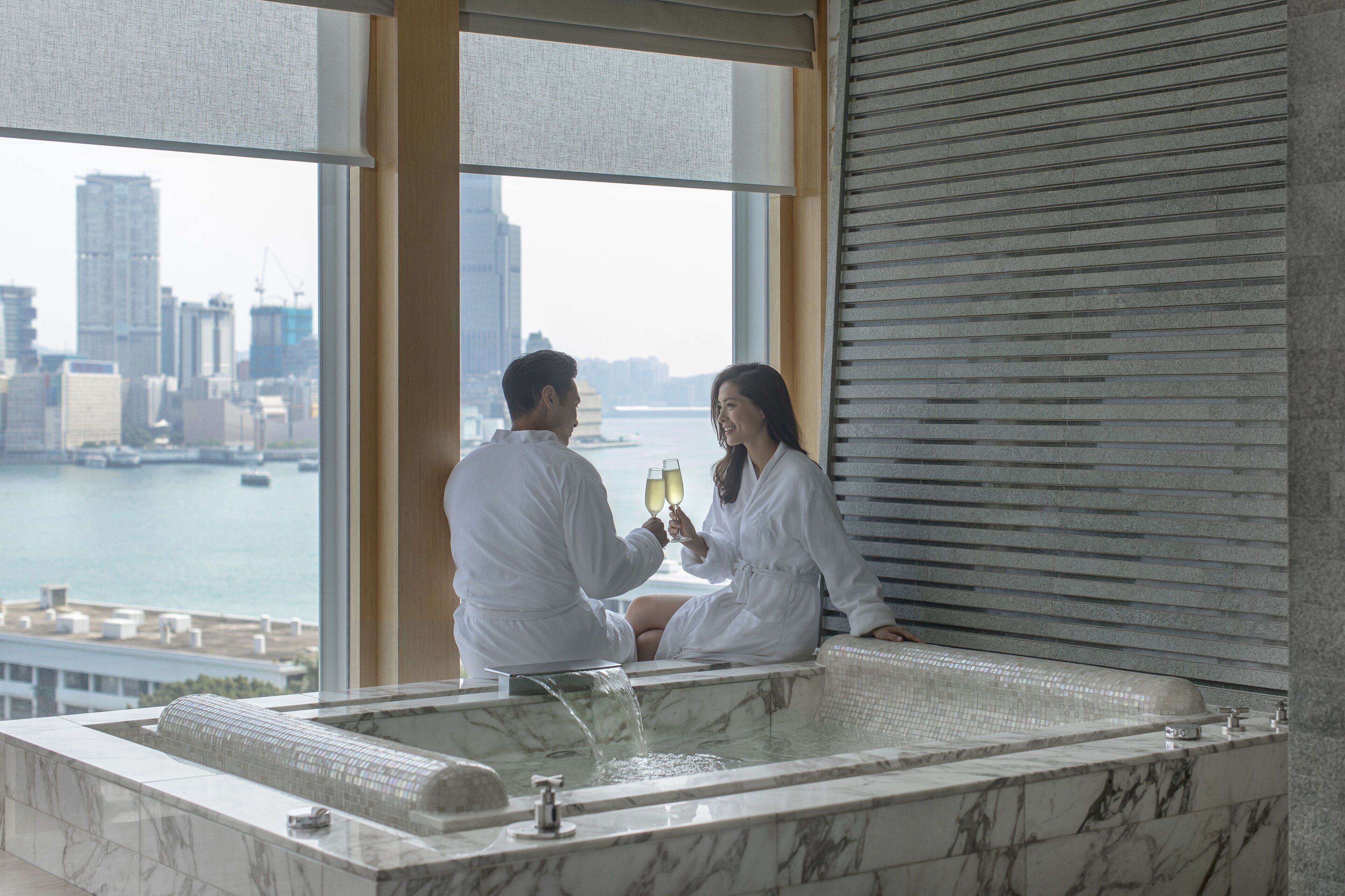 A couple in a bathtub with a city view, a large window, and a modern interior design.