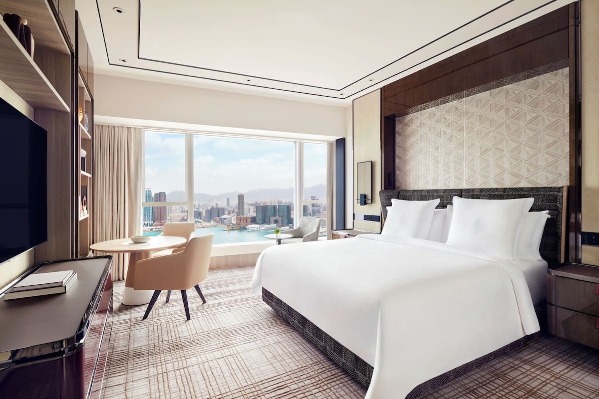 Een hotelkamer met een groot bed, een bureau, een stoel en uitzicht op een stadsgezicht door grote ramen.