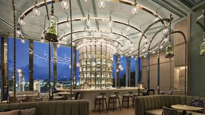 A bar area with a high ceiling, multiple hanging light fixtures, and a central bar stocked with bottles. There are seating areas with sofas and bar stools.