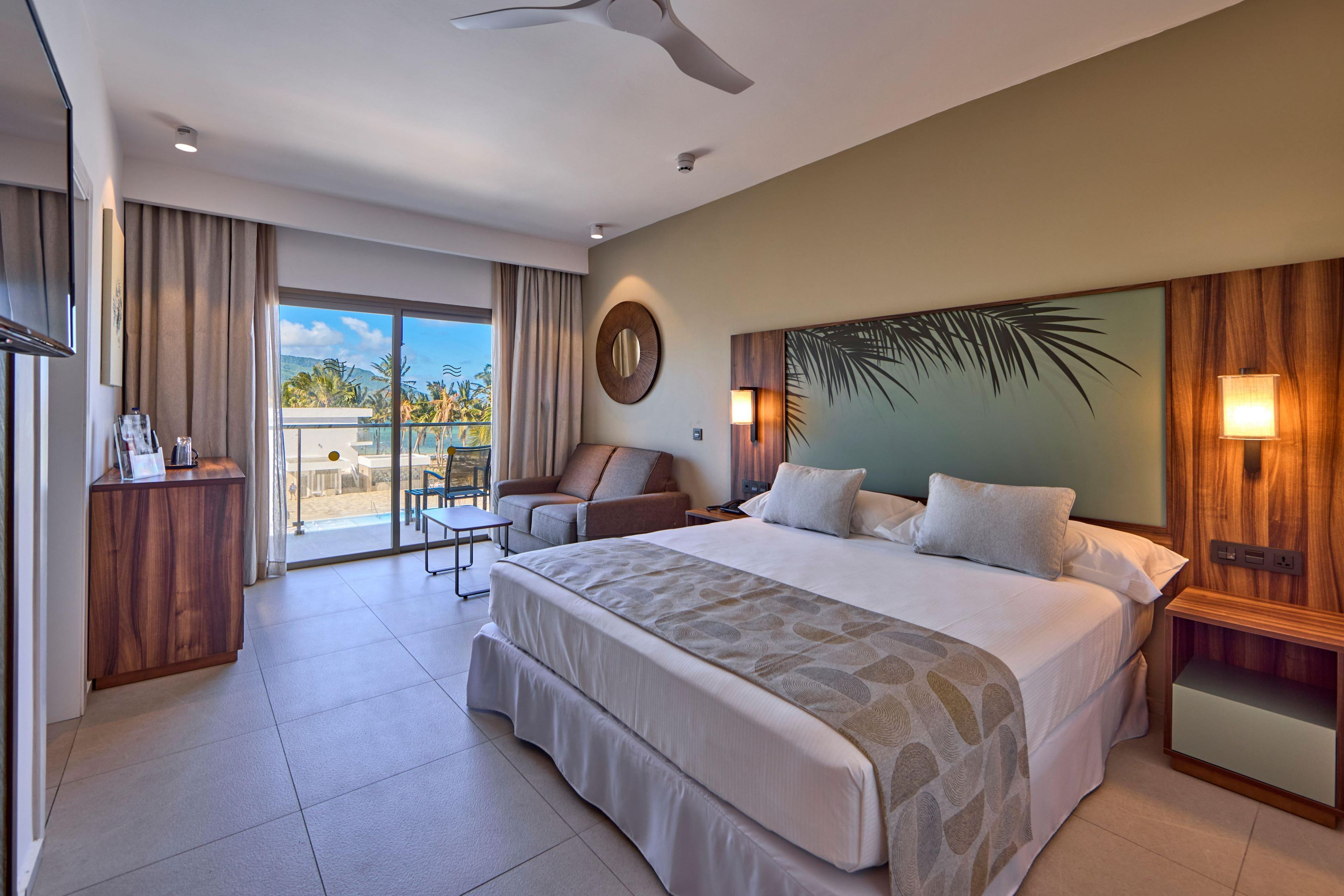 A hotel room with a large bed, a seating area, a balcony with a view, and a ceiling fan.