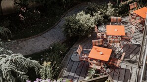 Eine Terrassengastronomie mit mehreren orangefarbenen Tischen und karierten Stühlen, umgeben von verschiedenen Pflanzen und Bäumen.
