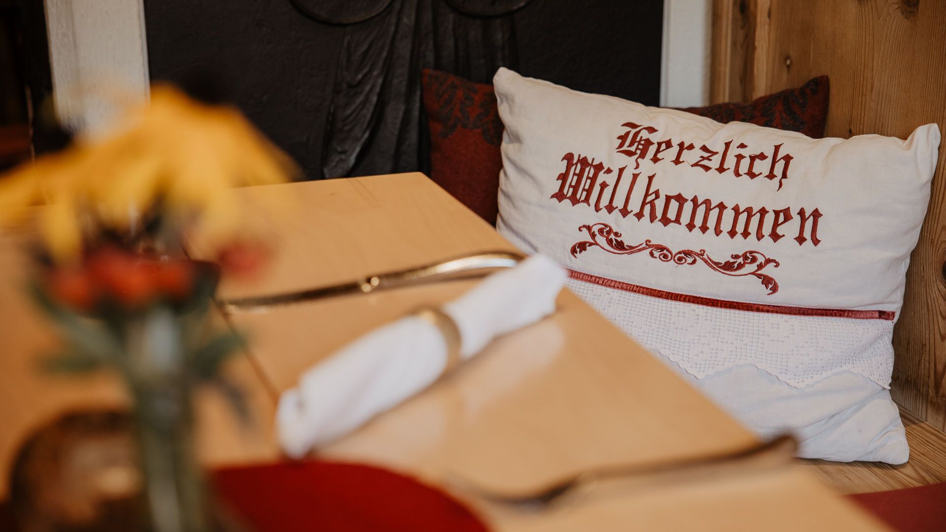Une table avec un coussin blanc sur lequel est inscrit le texte « Herzlich Willkommen » et un vase rempli de fleurs.