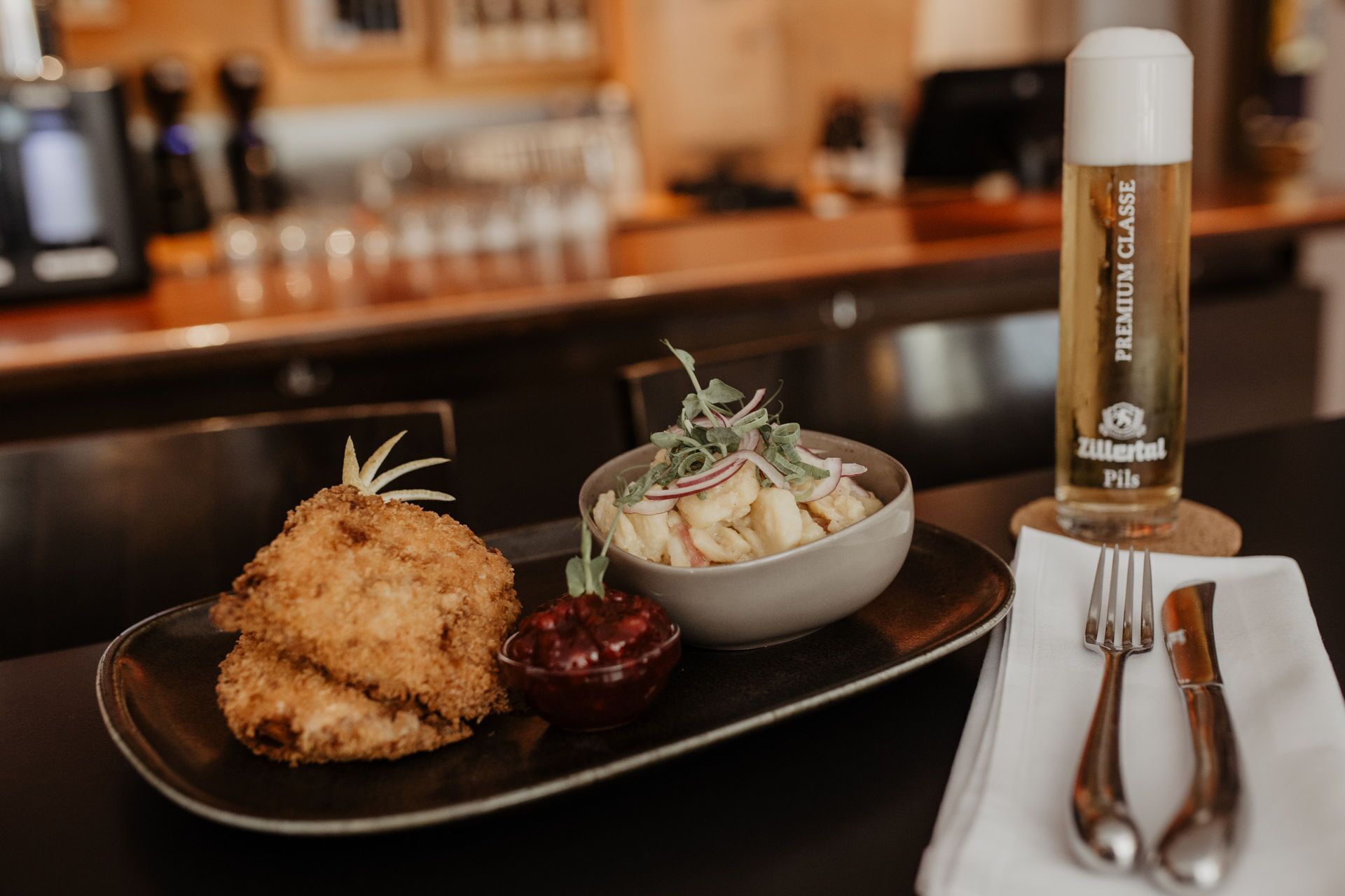 Un plato con un alimento empanizado, un bol de ensalada y una botella de cerveza Zillertai Pils sobre una bandeja.