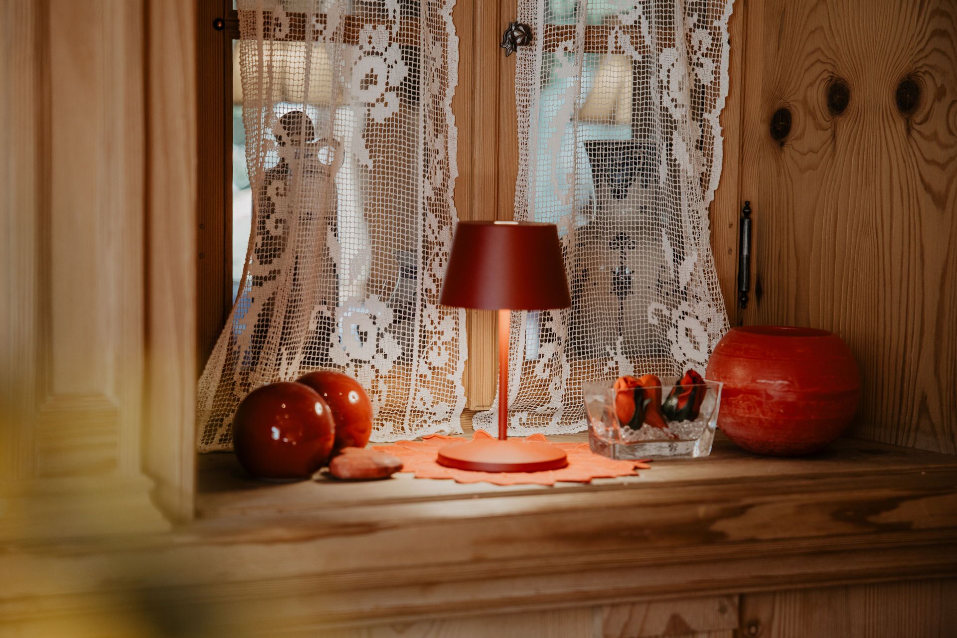 A wooden window sill with a lace curtain, a red lamp, a glass jar with a red object, and a pumpkin.