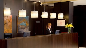 A reception desk with a dark wood finish, multiple pendant lights, and two abstract paintings on the wall. - NH Valladolid Bálago (Valladolid)