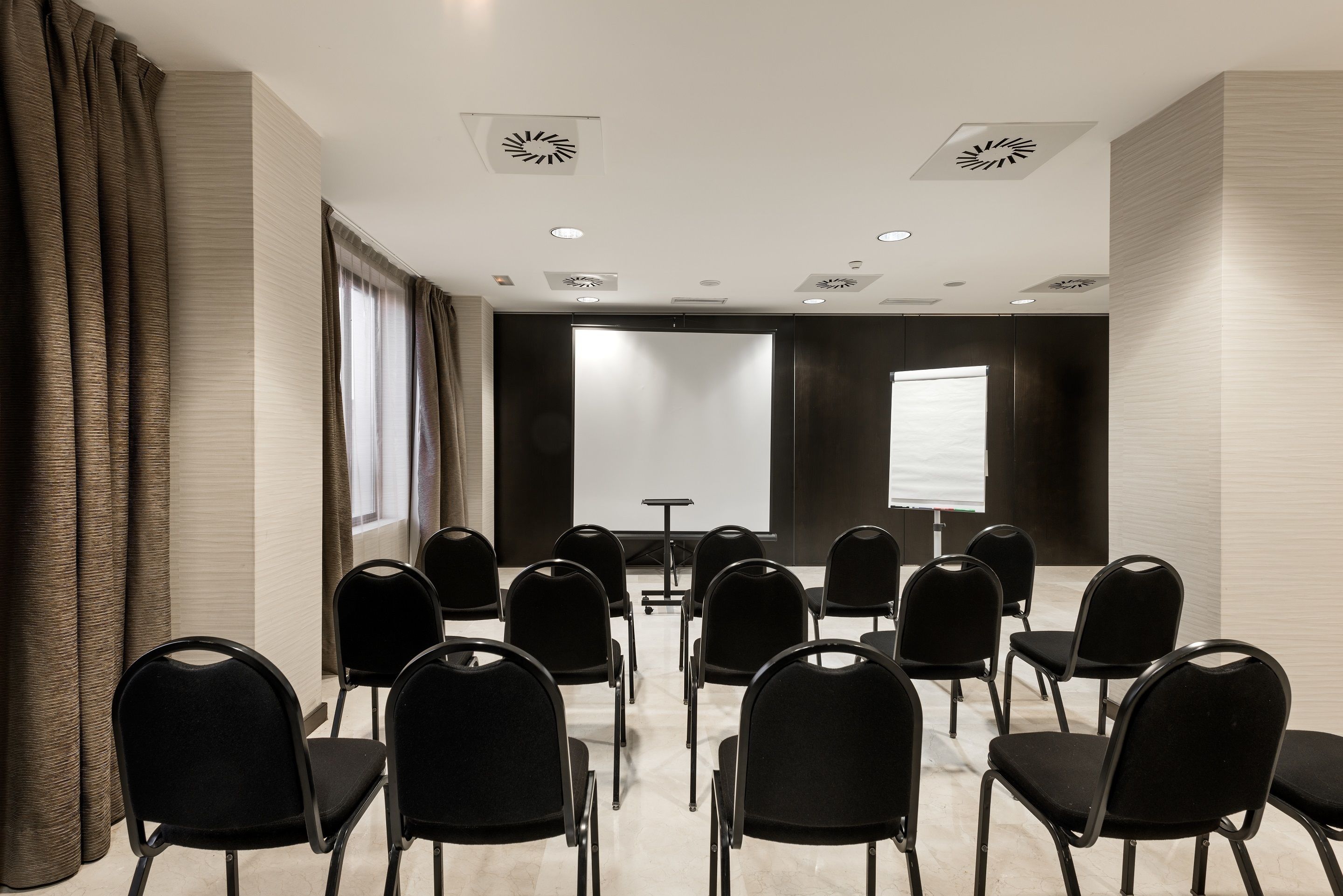A conference room with black chairs arranged in rows, a projector screen, and a table with a laptop.