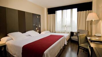 Habitación de hotel con dos camas, un escritorio, una silla, una lámpara y vistas a la ciudad a través de amplias ventanas.
