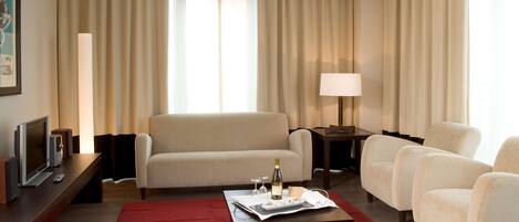 Habitación de hotel con sofá, sillones, una mesa de centro con una botella y vasos, televisor y lámpara.