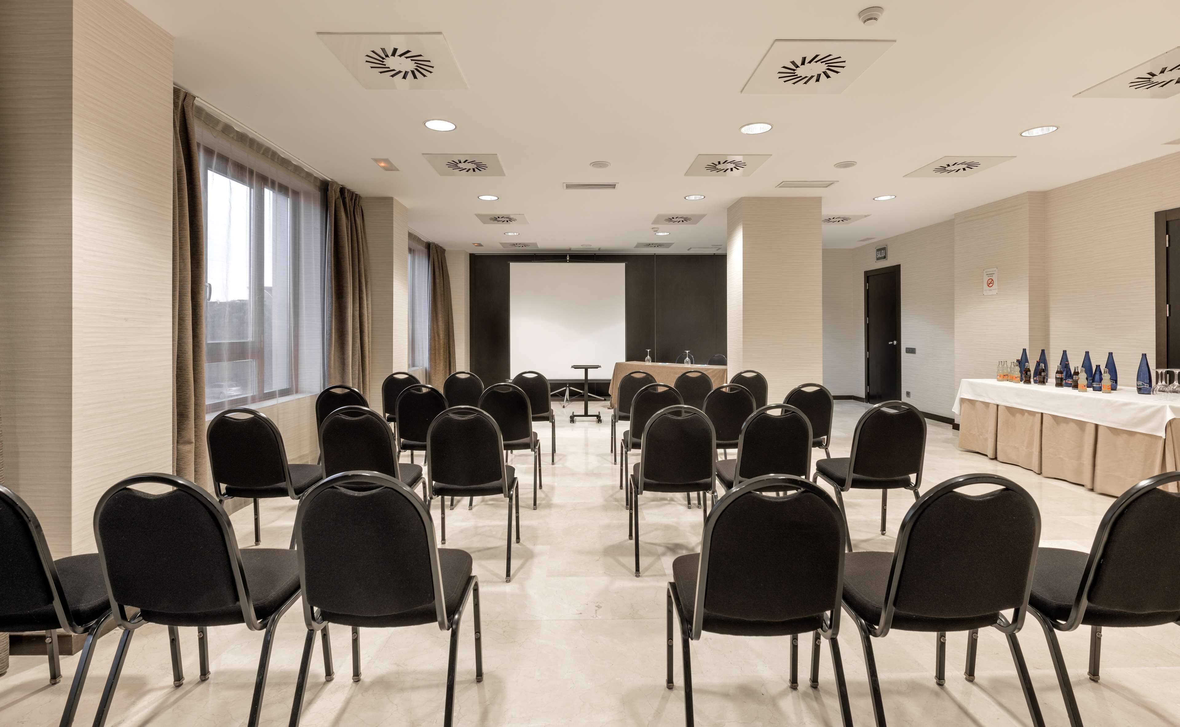 A conference room with rows of black chairs facing a long table with a projector screen at the front.