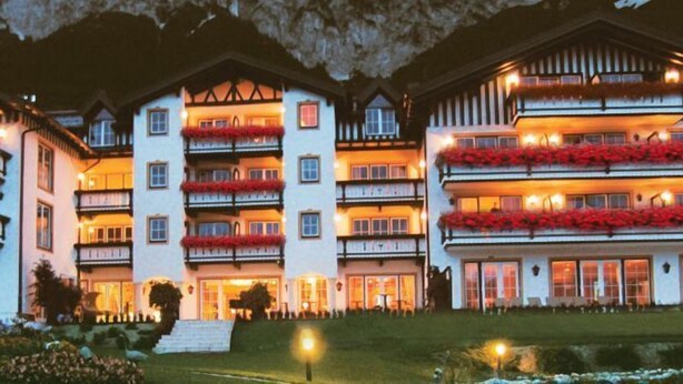 Un hotel de varios pisos con balcones decorados con flores rojas, una entrada bien iluminada y un paisaje montañoso de fondo.