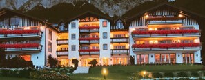 A multi-story hotel with balconies adorned with red flowers, a well-lit entrance, and a mountainous backdrop. - Alpenhotel Speckbacher Hof (Gnadenwald)
