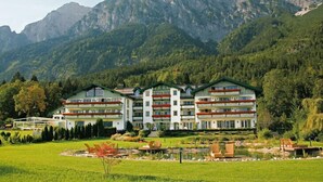 Un hotel de varios pisos con un jardín y un estanque al frente, rodeado de montañas.