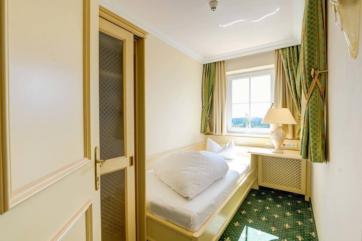 Una habitación de hotel con una cama, un ventanal con cortinas, una lámpara y una puerta con un panel de vidrio.