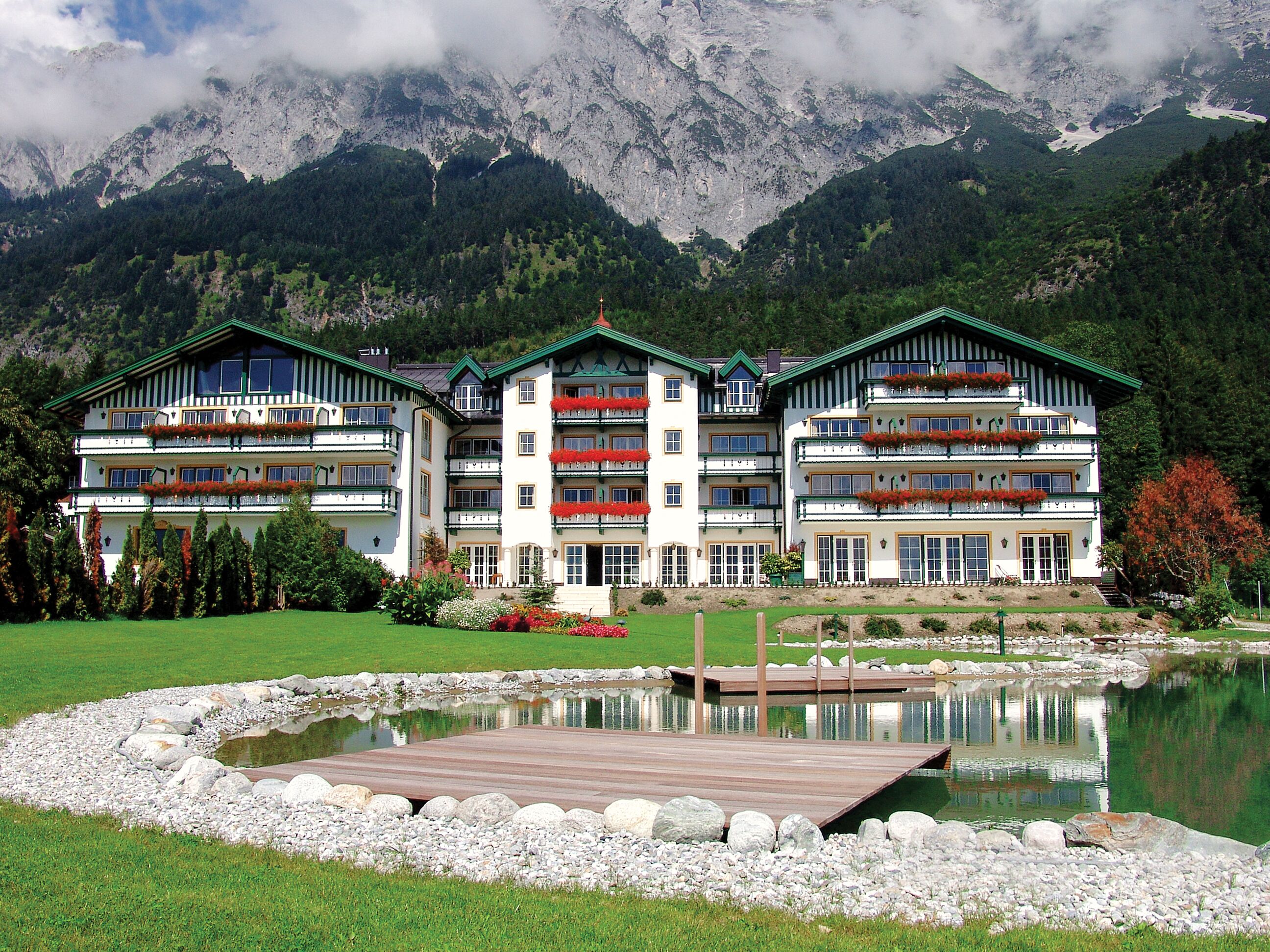 Un hotel grande de varios pisos con balcones decorados con flores rojas, situado junto a un lago con un muelle de madera, rodeado de montañas.