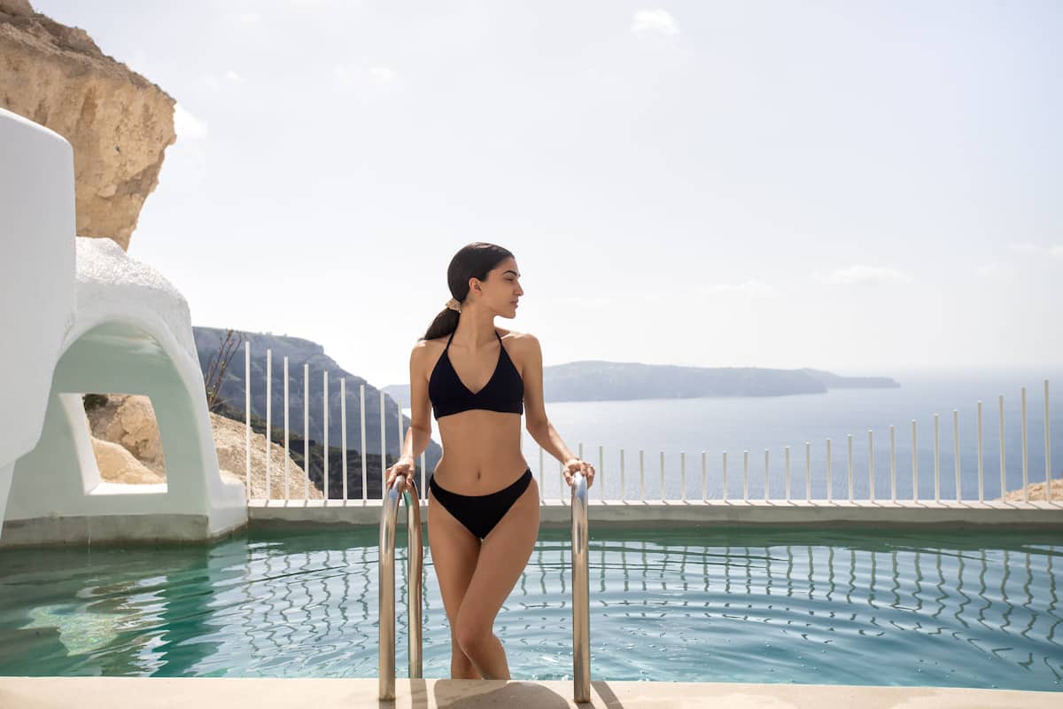 A woman in a bikini standing by a pool with a white fence and a view of the sea.