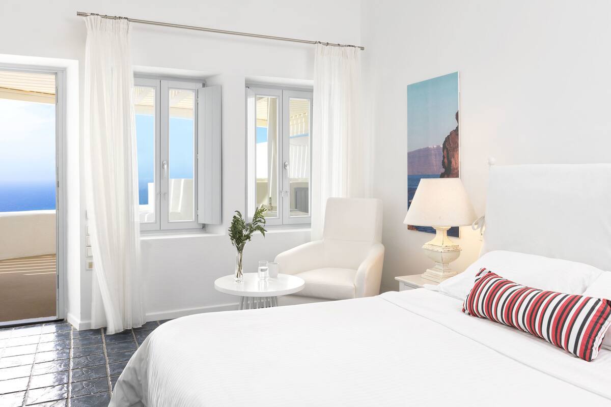 A hotel room with a bed, a chair, a small table with a vase, a lamp, and a painting on the wall.