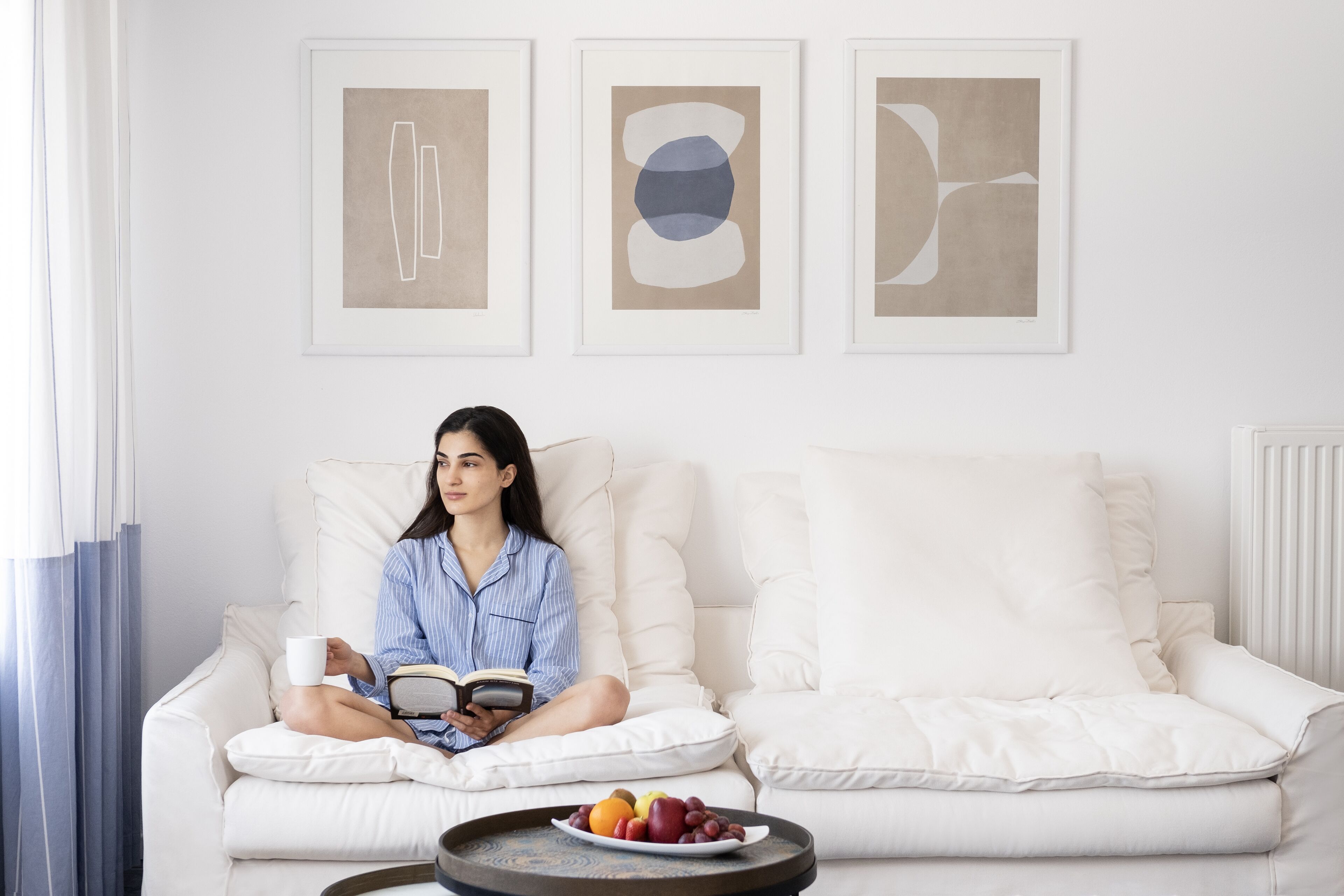 Uma pessoa sentada em um sofá branco, com uma bandeja de frutas à frente, lendo um livro.