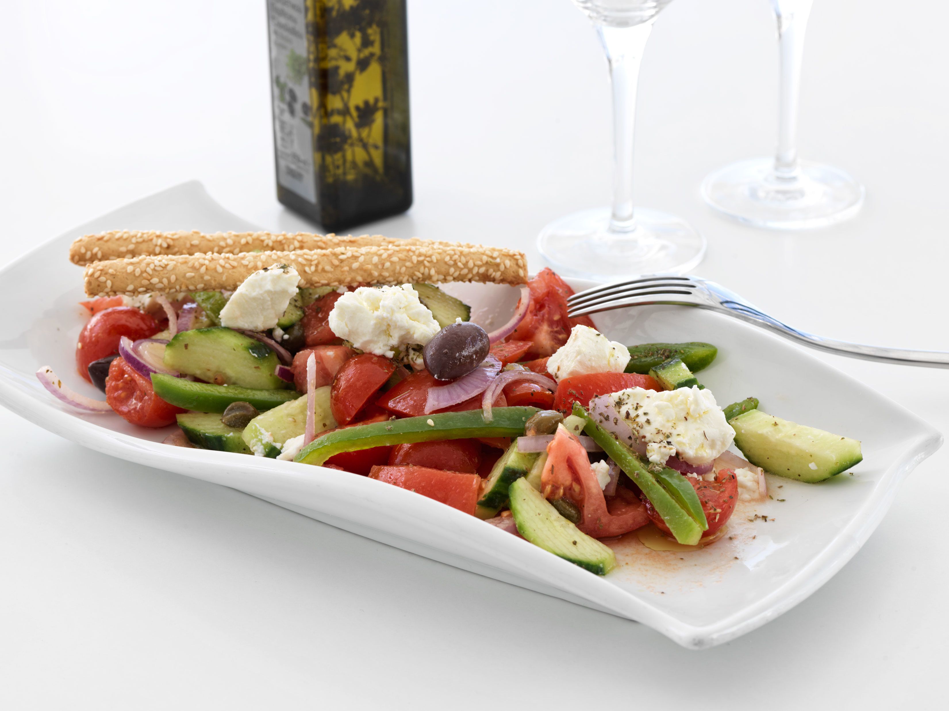 Een wit rechthoekig bord met een Griekse salade, twee crackers met sesamzaad, een glas witte wijn en een fles olijfolie.