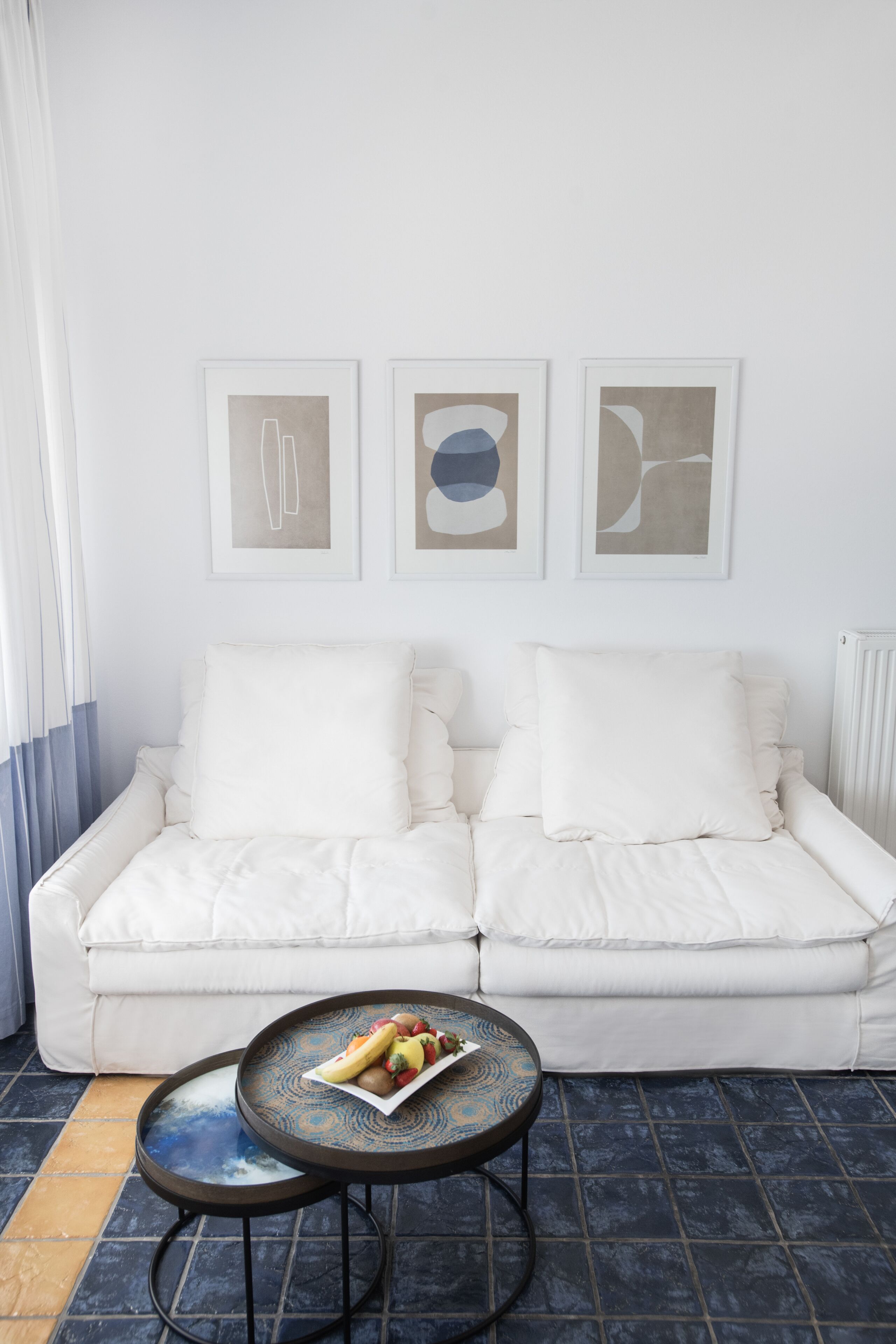 A modern living room with a white sofa, a round coffee table with a fruit bowl, and abstract wall art.