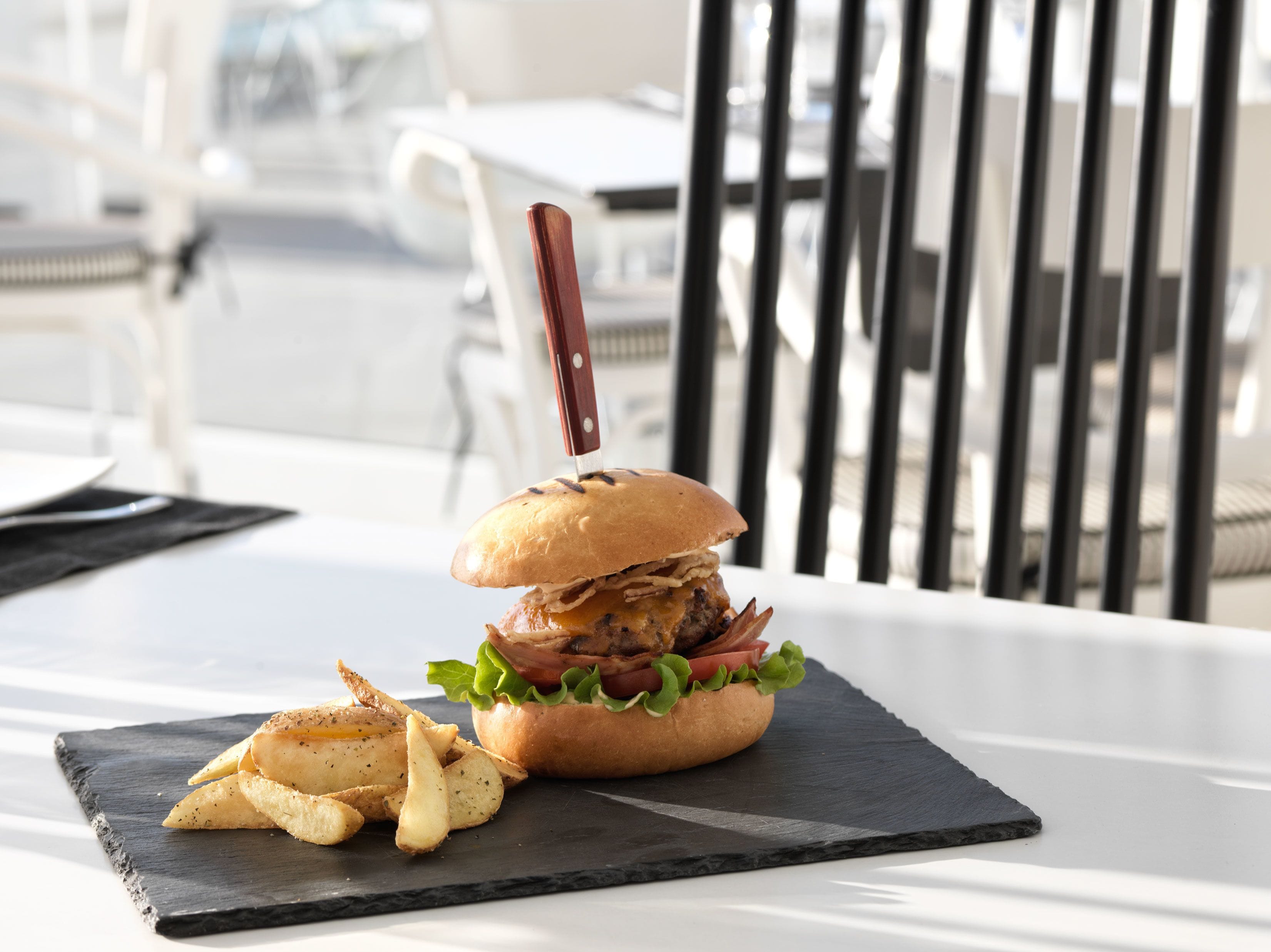Ein Burger mit Messer, serviert auf einer schwarzen Schieferplatte mit Kartoffelspalten.