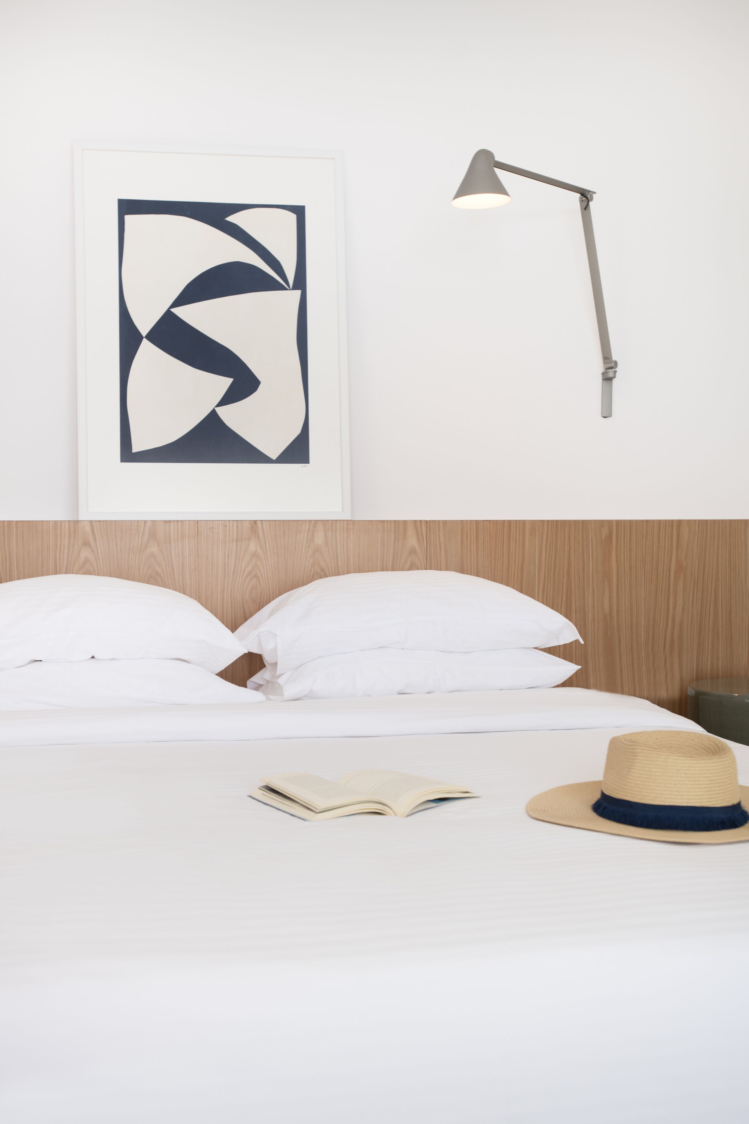 A neatly made bed with white linens, a single open book, and a straw hat on the bed. A modern desk lamp is mounted on the wall to the right.