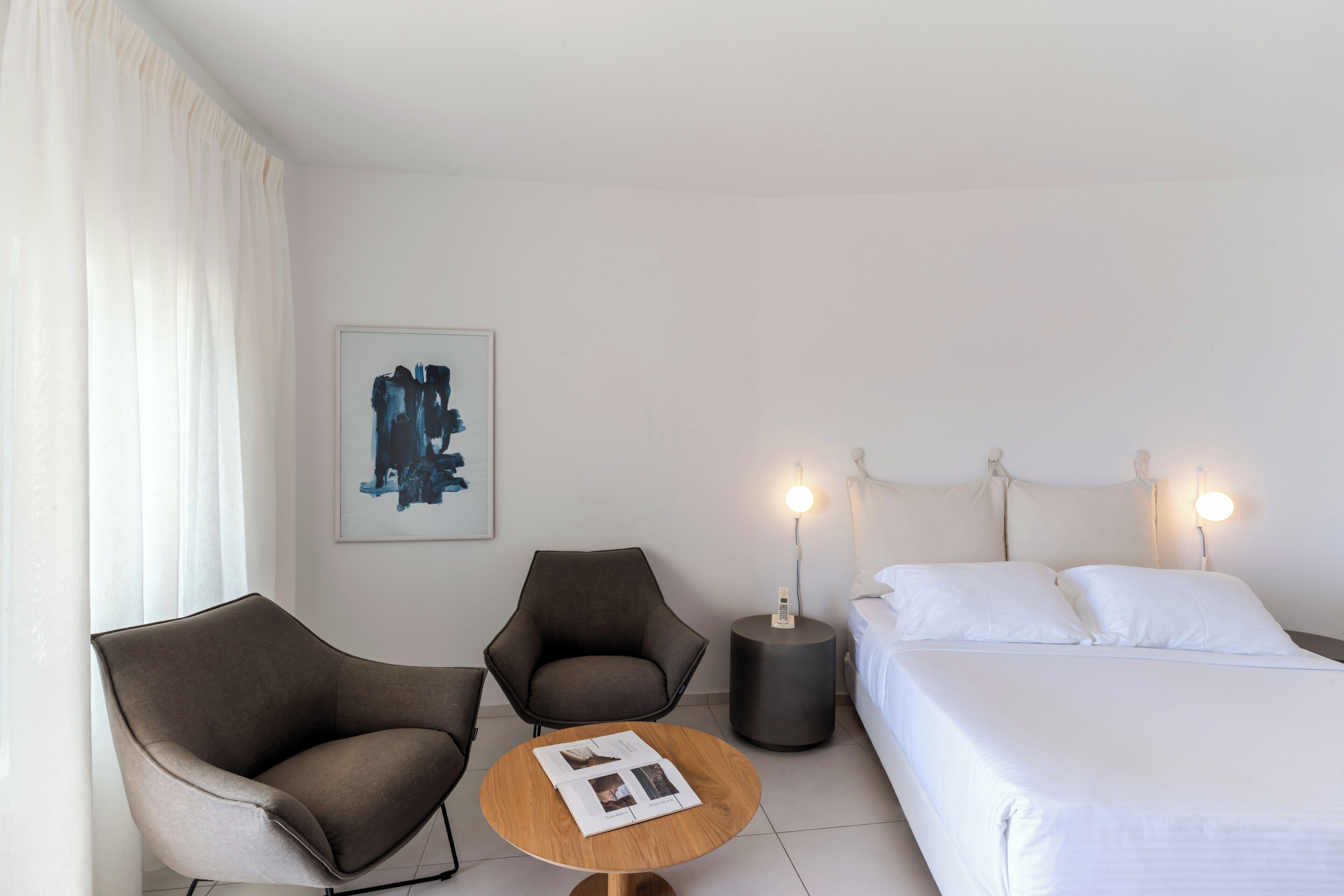 Une chambre d’hôtel comprenant un lit, deux fauteuils, une table ronde avec des magazines, une lampe de chevet et une œuvre d’art murale.