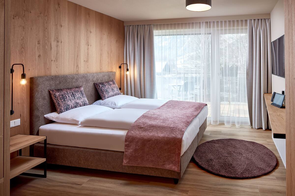 A modern bedroom with a wooden headboard, a large bed with a pink throw, a round rug, and a view of a balcony through sheer curtains.
