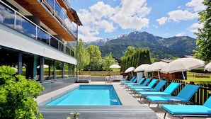 A modern pool area with blue lounge chairs and umbrellas, surrounded by lush greenery and a mountain backdrop. - Hotel Sonnblick (Kaprun)